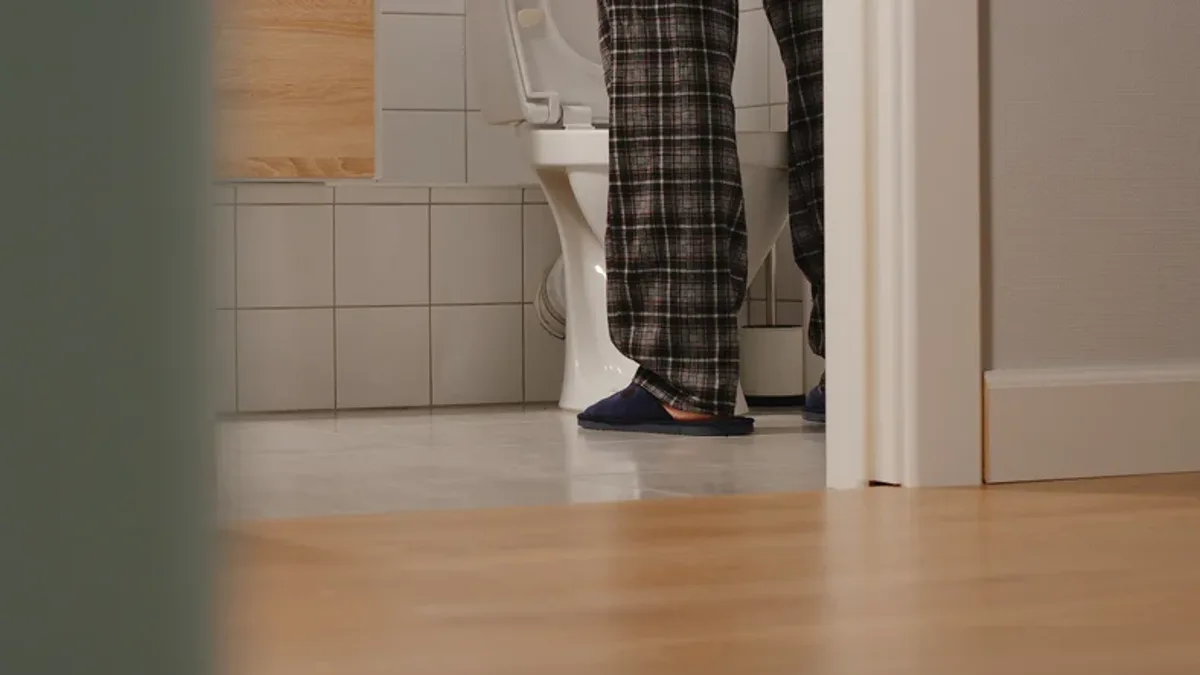 A man's legs and slippers in a dark hallway, walking towards a lit bathroom at night